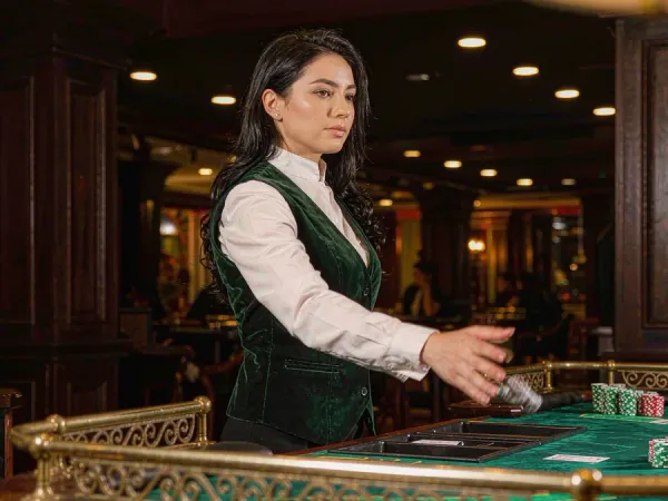 m66 Dealer in a green velvet waistcoat reveals cards at a baccarat table with chip stacks